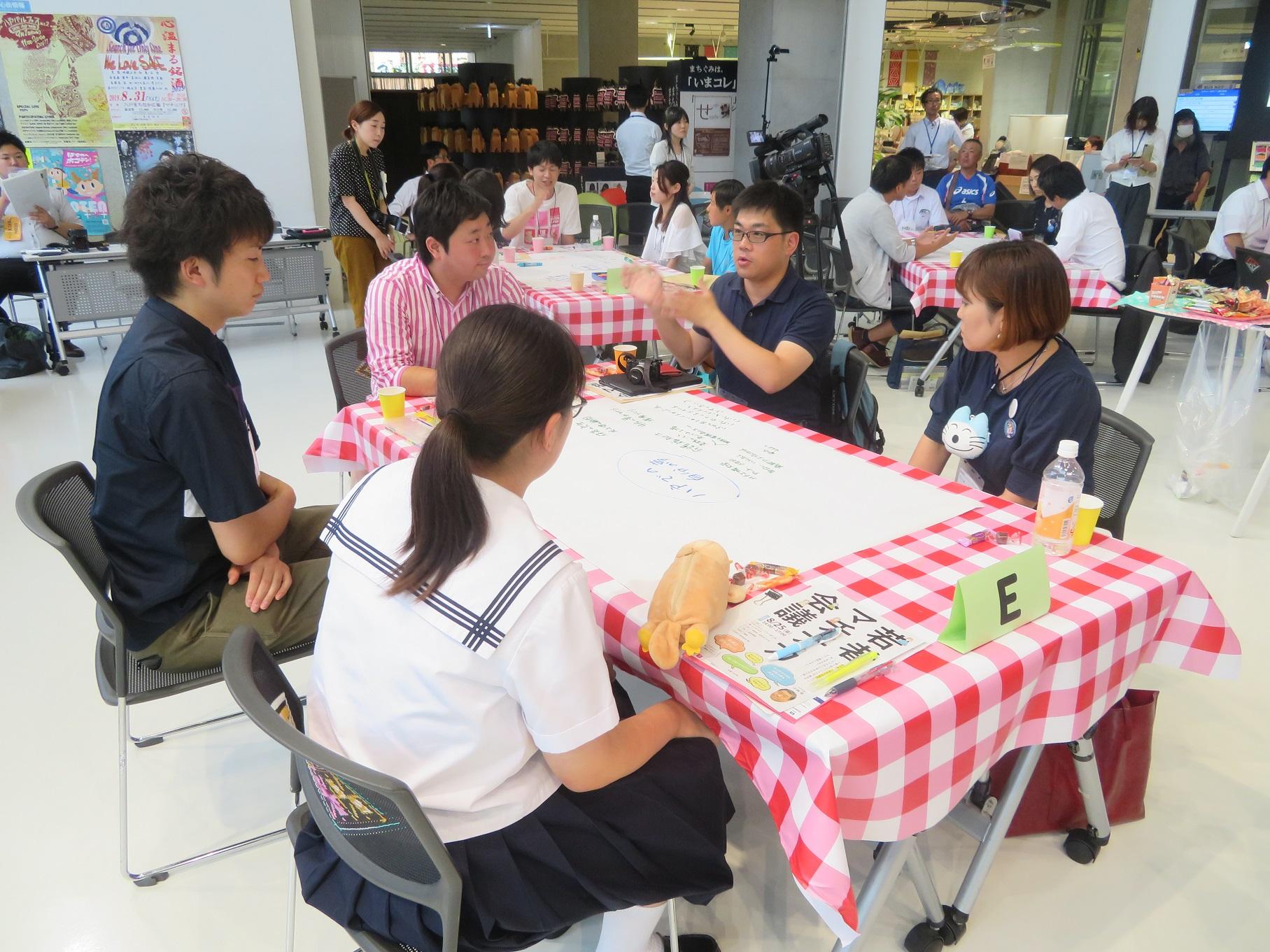 5名の男女がテーブルを囲んで座って話をしている写真（手前に制服を着た女性）