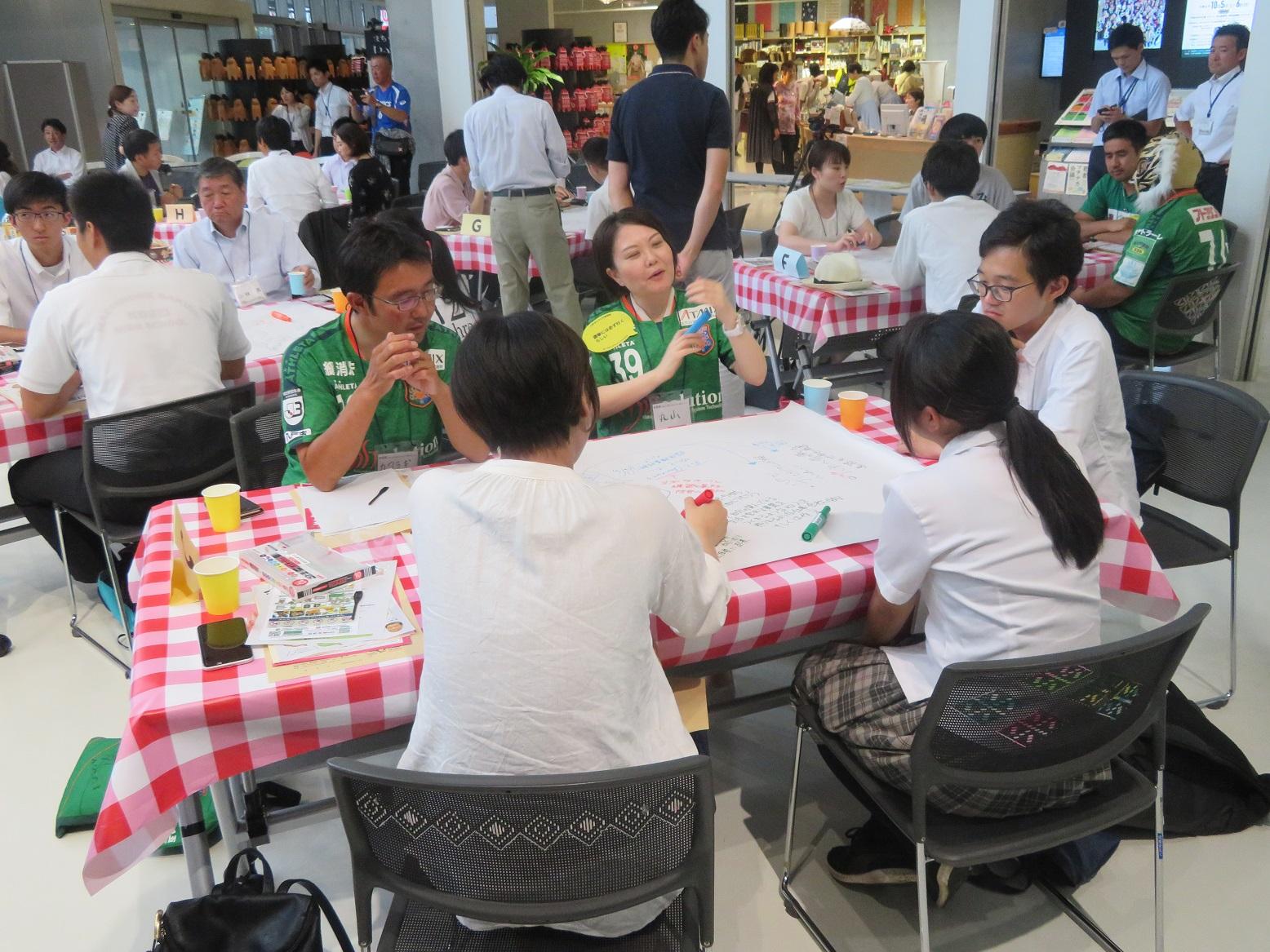 テーブルを囲んで座って話をしている男女5名の写真（テーブル奥側に緑の服を着た男女2名）