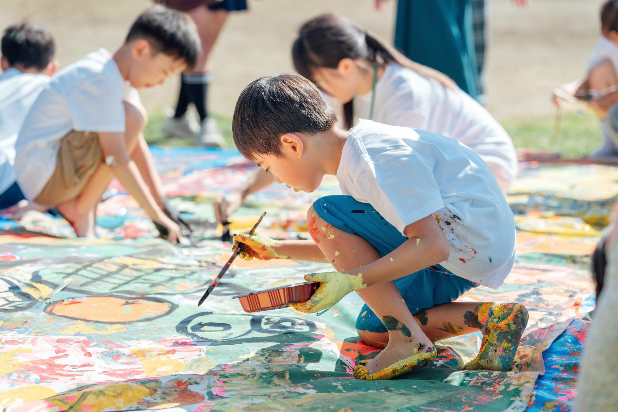 子どもたちが広いキャンパスに絵を描いている風景。男の子が真剣な表情で筆を持ち、鮮やかな色の絵具で何かを描いている。