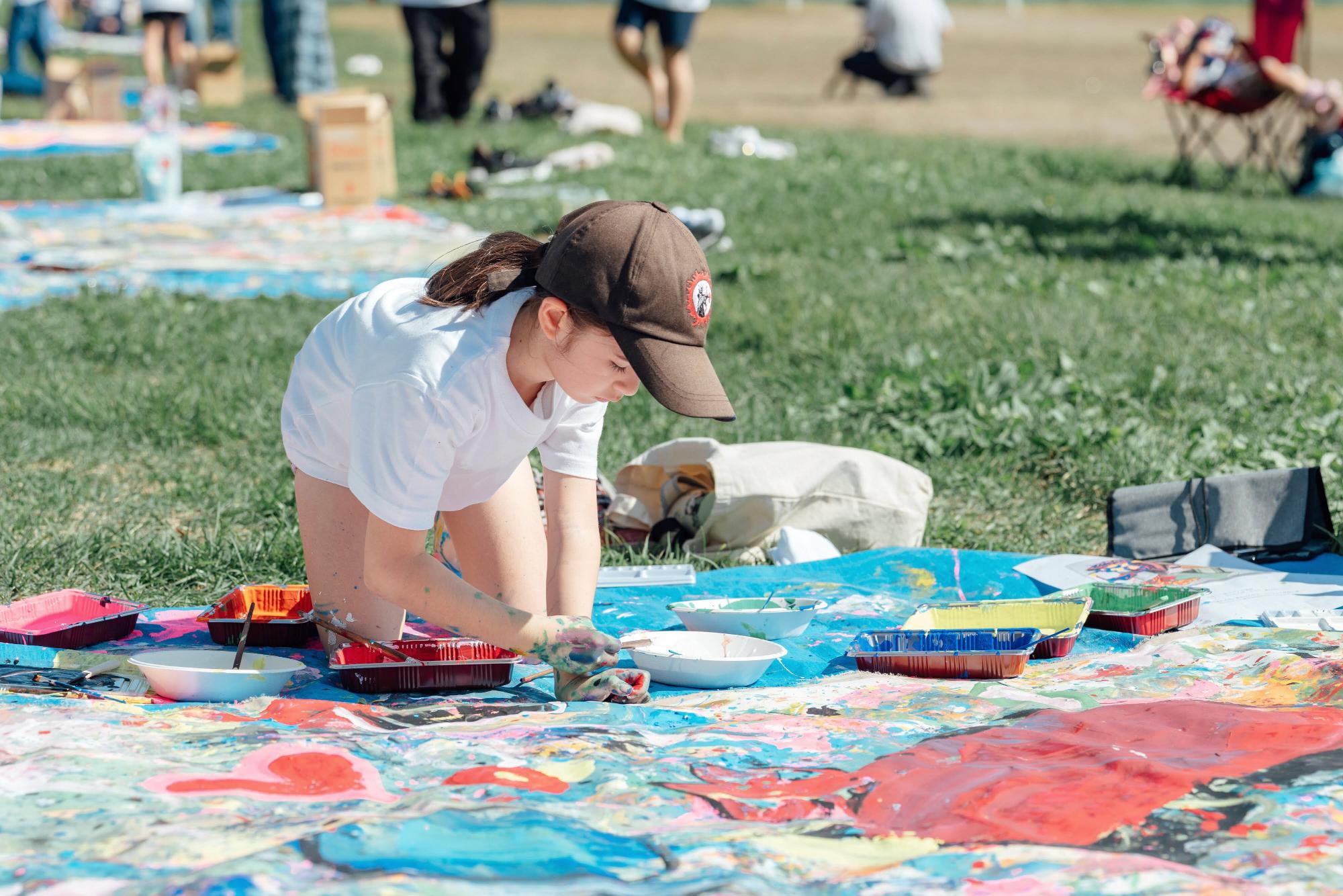 白いTシャツを着た女の子が、色とりどりの絵具で地面の広げたキャンパスに手を付けて作業中。その周囲には他の子どもたちも楽しそうに活動している。