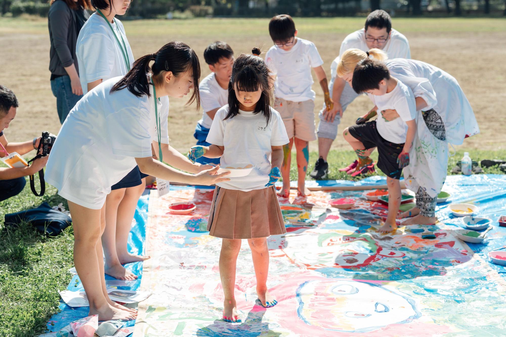 女の子が他の子どもたちと一緒に大きなアートシートの上で絵を描いている様子。周りにはカラフルな絵の具が散らばっている。