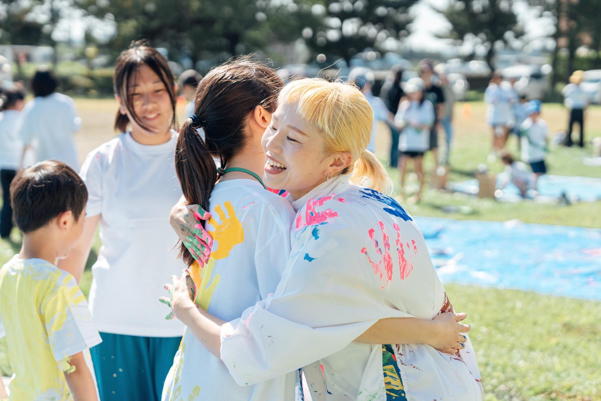 参加者の女子と講師が笑顔で抱き合い、互いの白シャツに絵具をつけ合って楽しんでいる様子。