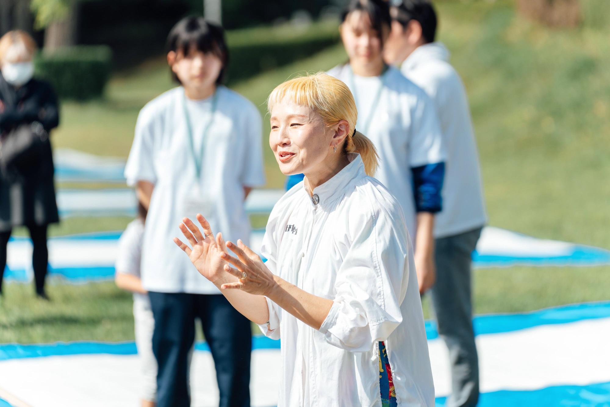 白い服を着た講師が、参加者に向けて話しかけている場面。