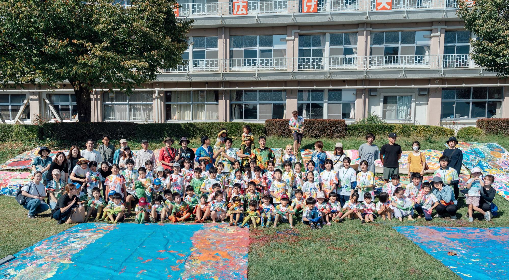 大勢の参加者たちが色とりどりのアート作品の前に集まる様子。背景には学校の建物と緑の草地が広がっている。