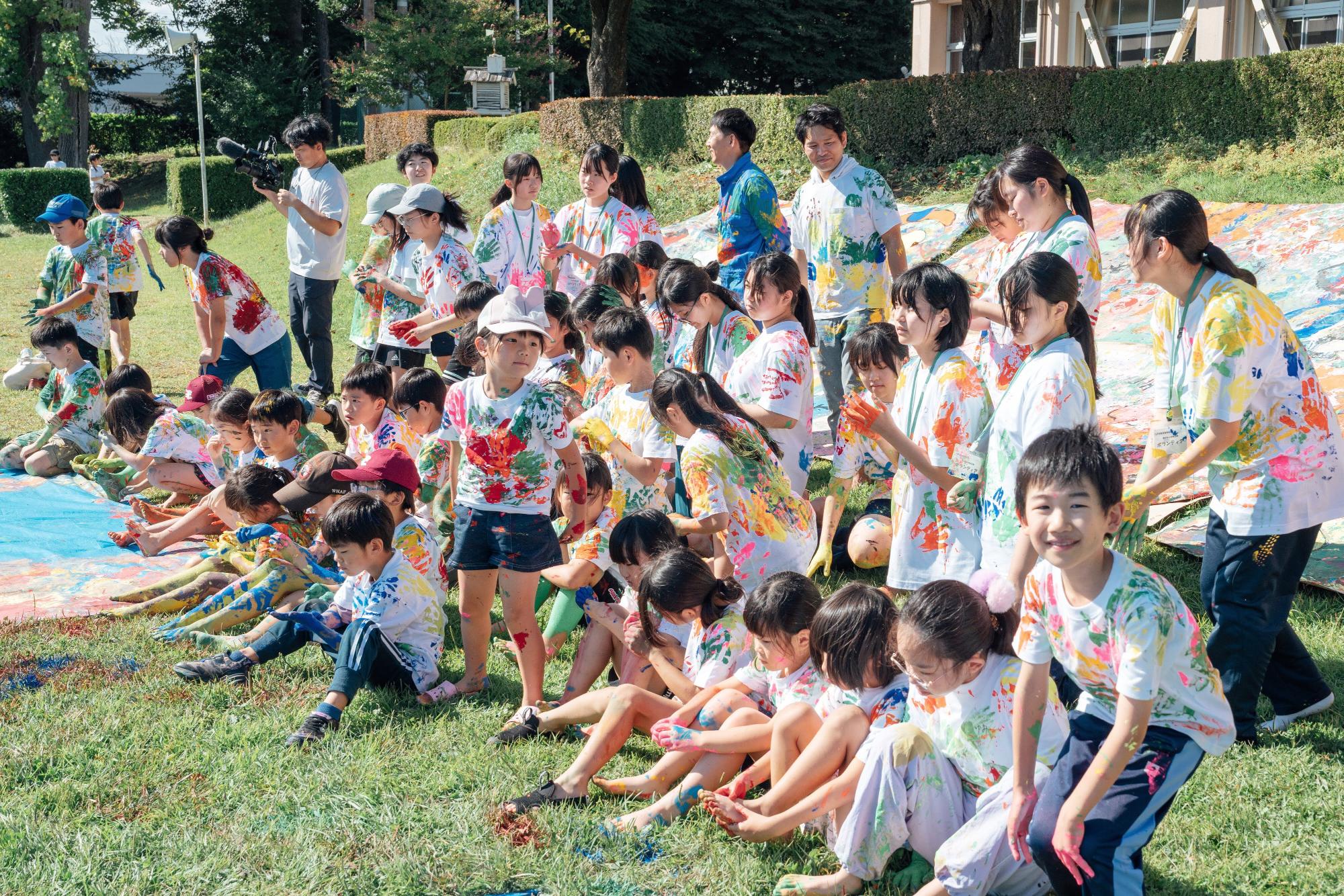 カラフルな絵具のついたTシャツを着た子供たちが、広い草地に笑顔で集まっている。