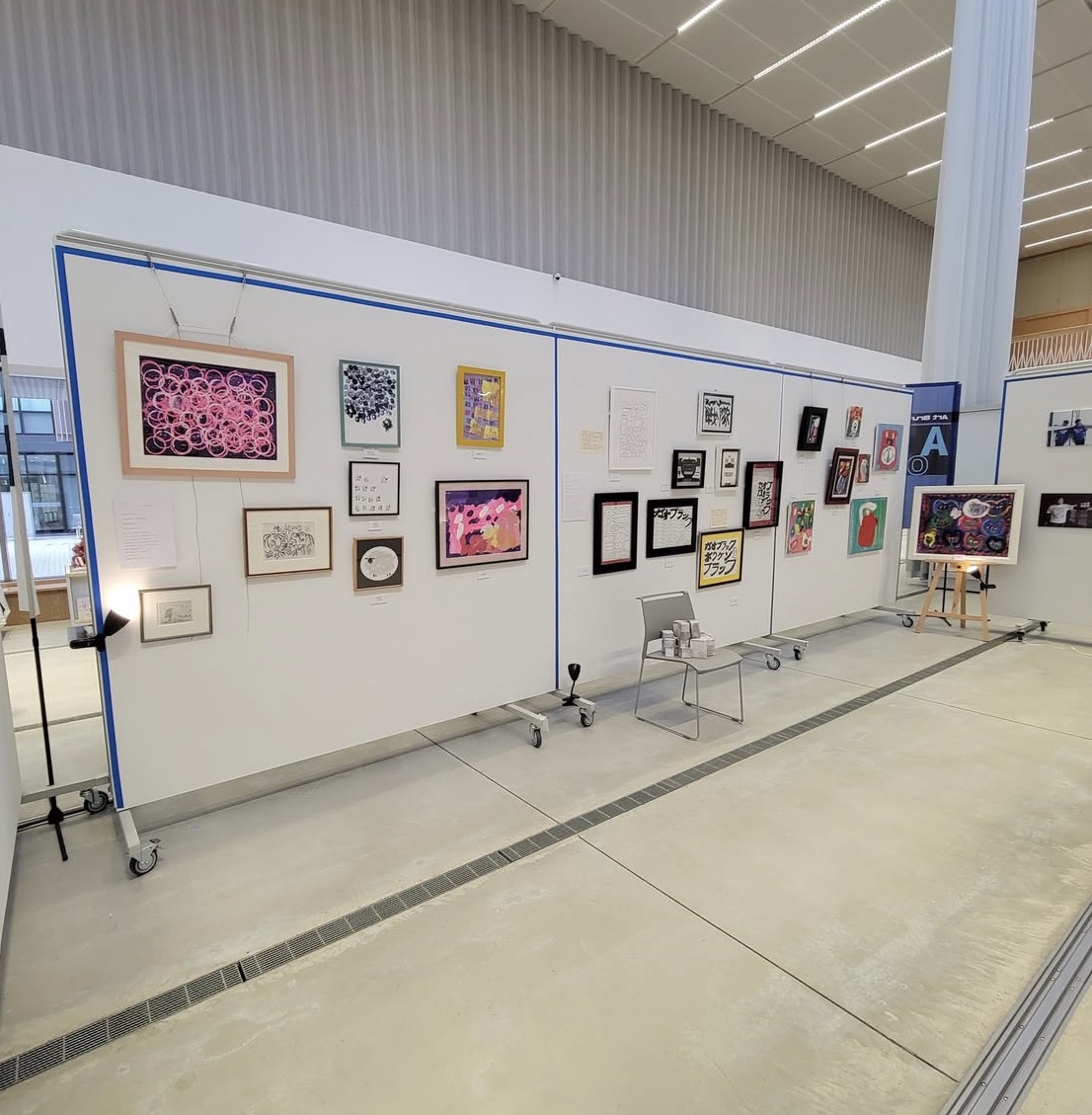 展示会の壁に飾られたアート作品の数々。様々な形や大きさのフレームが見られ、中央には椅子が配置されている。