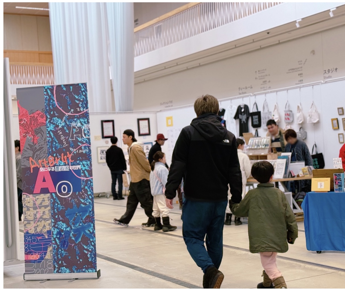 複数の人々がアート展示会を訪れている様子。手をつないで歩いている親子が見え、展示品が背景にならんでいる。