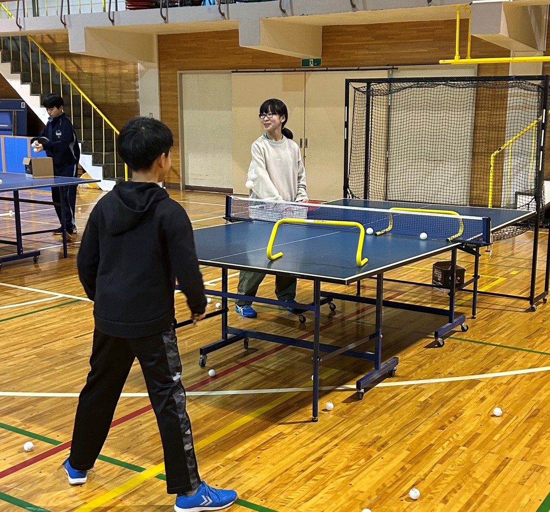 子どもたちが卓球の練習を楽しんでいる光景。ネットを挟んで対戦しながら、楽しそうにプレイする姿が描かれている。