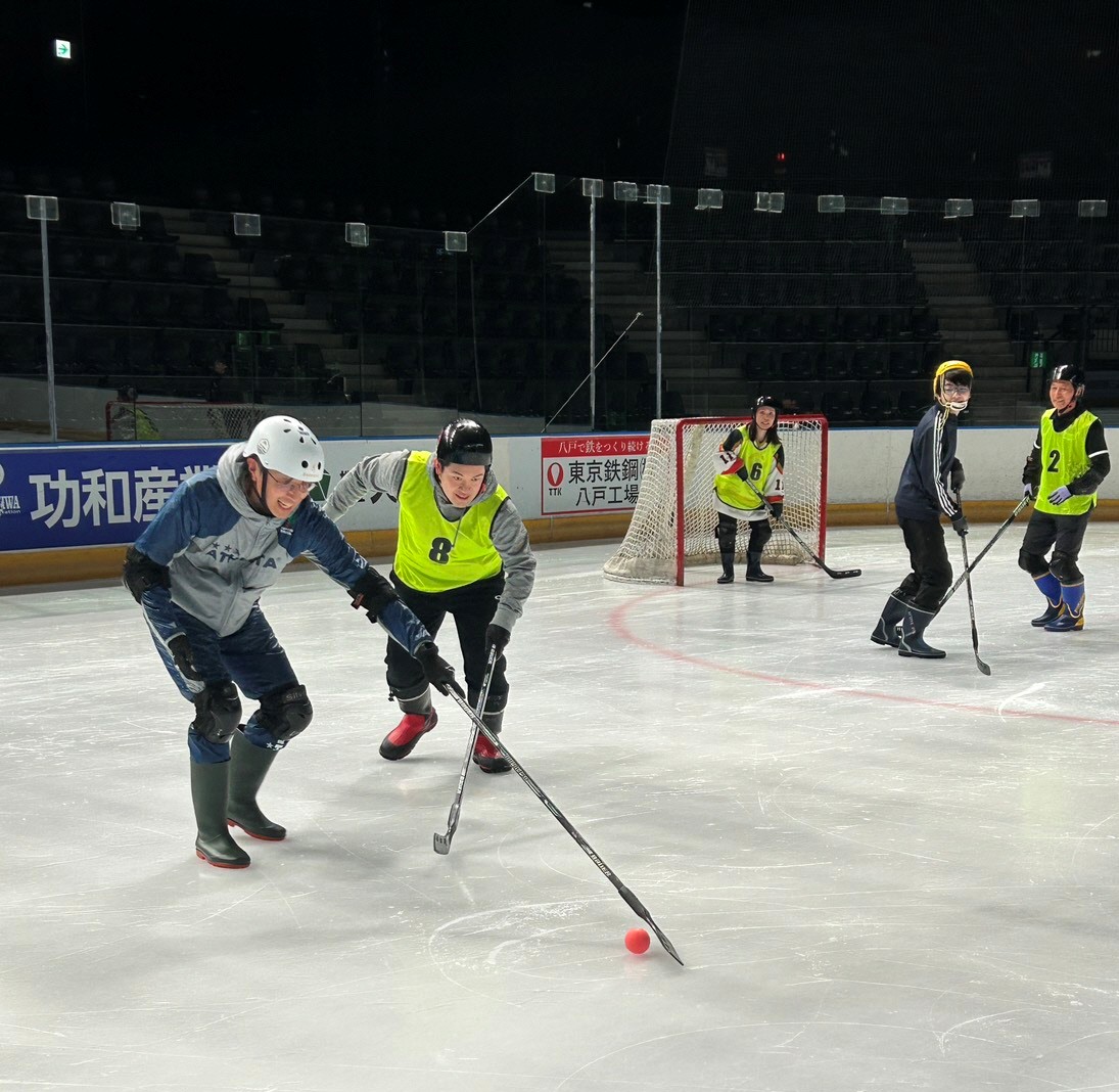 長靴アイスホッケーをプレイする選手たち。黄色のビブスを着用した選手が、パックを追いかけている。氷の上にはほかの選手も見られ、競技の雰囲気が感じられる。