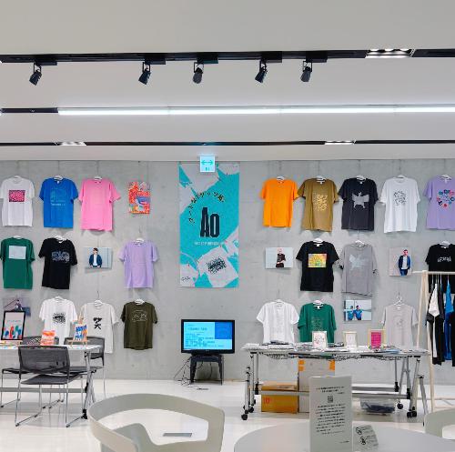展示スペースに並んだ様々ないⓇののTシャツが壁にかけられている様子。中央に「アートなTシャツ展」のポスターが掲示されている。