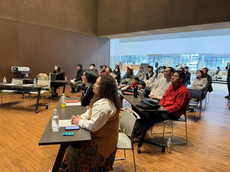 講演を聴く来場者たち。会場前方でスピーカーの説明を受け、参加者が座ってメモや視聴中。