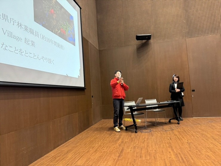 会場でプレゼンテーションを行う人物とスライドが映った檀上の様子