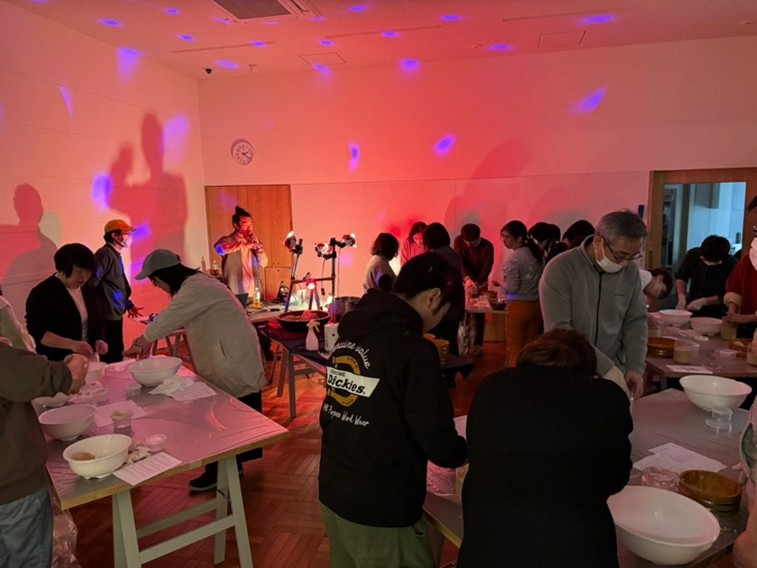 独特な赤い照明の室内でみそ仕込みの実習をする参加者たちの様子。テーブル上にボウルが並び、数名が作業中。