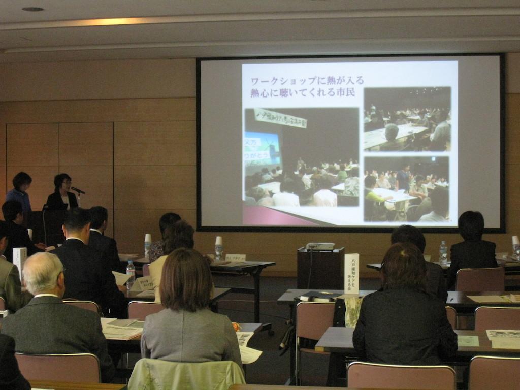会議室でスクリーンに映し出される事業のスライドを見る審査員を会場後方から写した写真