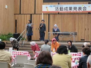 エプロンと頭巾をした3人の女声が壇上でわらべ歌を歌っている写真
