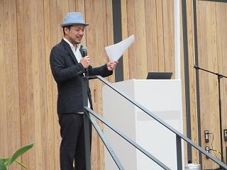 ステージ上の演壇で事業内容を発表するグレーのハットをかぶり黒いスーツを着た男性の写真