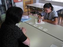 外国語を母国語とする女生徒が机に向かいノートに記入している向かい側から講師の女性が指導をおこなっている写真。
