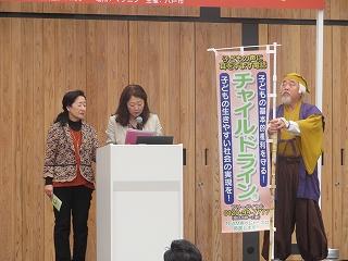ステージ上で事業内容を発表する水戸黄門の衣装をきた男性と二人の女声のの写真