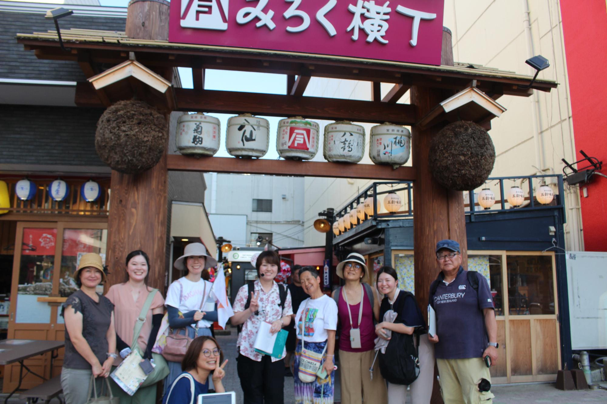 みろく横丁の看板と酒樽提灯が見える木製門前で、八戸ディスカバリーガイド養成講座実施委員会の会員と受講生メンバー合わせて9名が記念撮影