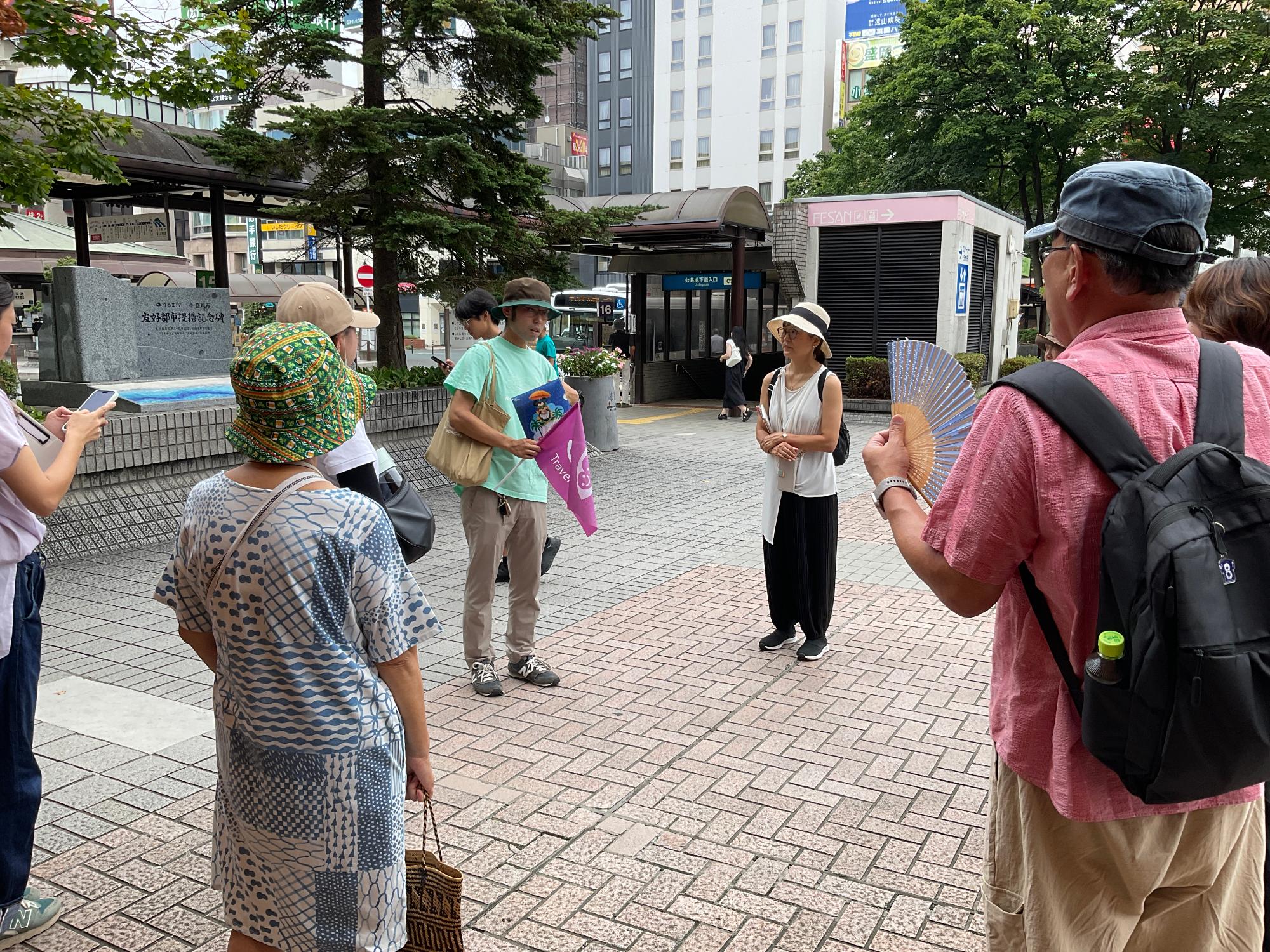 街中の広場で旗を掲げるガイドが説明をする中、参加者が地図を手に聞き入る場面