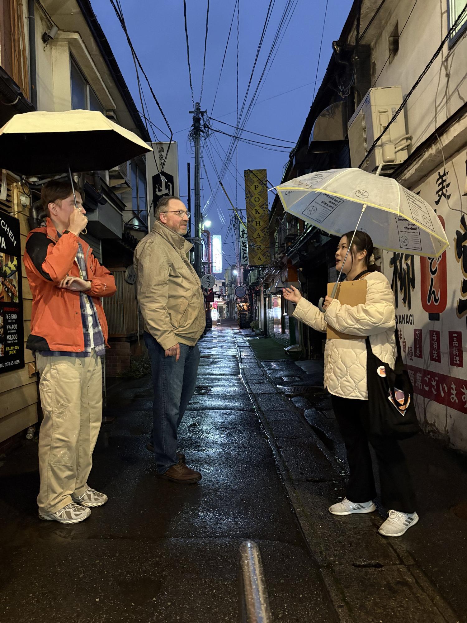 夜の路地で傘をさして話す3人、濡れた路面と電線が見える