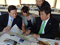食堂のテーブルで食事をしながら資料を見る二人の市長と地図を指差し説明している女性の写真。