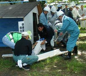 白い布で包んで担架に乗せようとしている人や周りにヘルメットをかぶった人たちが大勢いる救出訓練中の写真