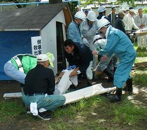 白い布で包んで担架に乗せようとしている人や周りにヘルメットをかぶった人たちが大勢いる救出訓練中の写真