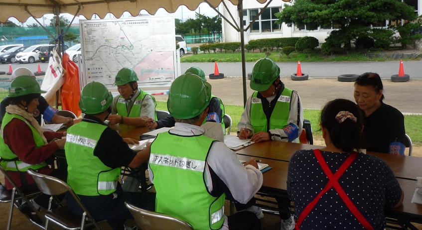 緑のセーフティベストとヘルメットを身に付けた人たちがテントでミーティングをしている写真