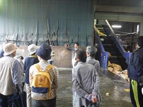 リサイクル施設の中でプレス機の説明をしている北日本産業株式会社社員と説明を聞いているエコツアー参加者の写真