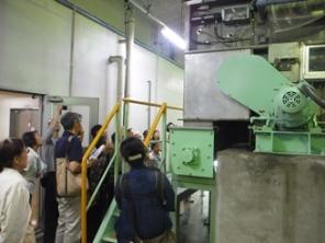 工場内の脱水機の機械の前で、見学者が説明を聞いている様子の写真
