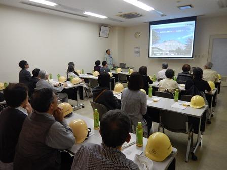 会議室にて施設の見学者が席に着いており、机には黄色いヘルメットが置かれ、前方の映像を見ながら事業概要の説明を聞いている様子の写真