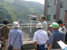 ダムの上で写真を手に持ち説明している男性と、話を聞いている参加者たちの写真
