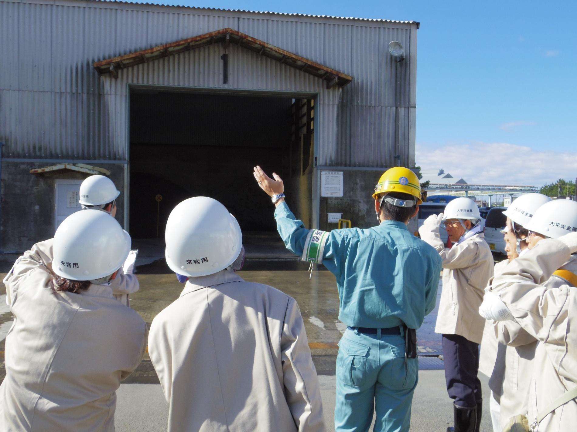 倉庫のような建物の一般廃棄物置き場で、黄色いヘルメットを被り左手を挙げて説明をしている大平洋金属株式会社社員と話を聞いているエコツアー参加者の写真