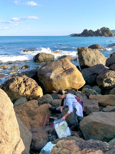 波打ち際の大きな岩の間のごみをトングで拾う女性の写真