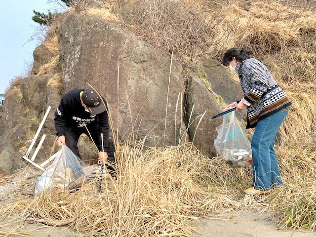 大きな岩の前で枯れた草の中からトングでゴミを拾う二人の写真