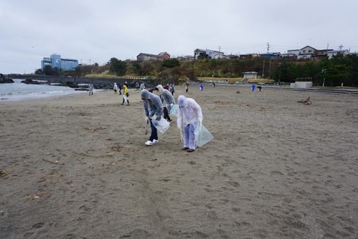 雨の中雨合羽を着て砂浜のごみを拾う参加者の写真