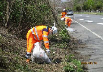 オレンジ色の作業服を着て道路わきの木々の多い中、ごみを集め袋に詰める人の写真