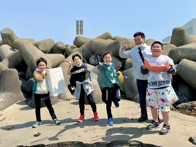 砂浜のテトラポットの前で拾ったごみを手にした子供たちの写真