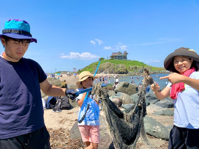 蕪嶋神社を背景に、砂浜にあった漁網をトングで三人で持ち上げている写真