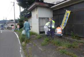 道路脇の草の生えた場所のごみを拾う写真