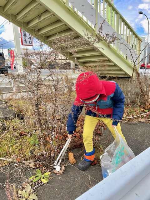 ボランティア用ごみ袋とトングを両手に持ち、歩道橋下のごみを拾っている子供の写真