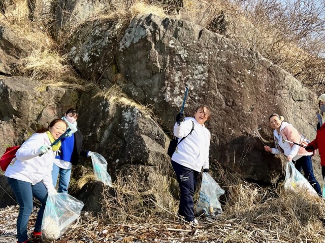 枯草の生えた大きな岩のある場所でごみ袋とトングを手に、ごみを拾う数人の女性の写真