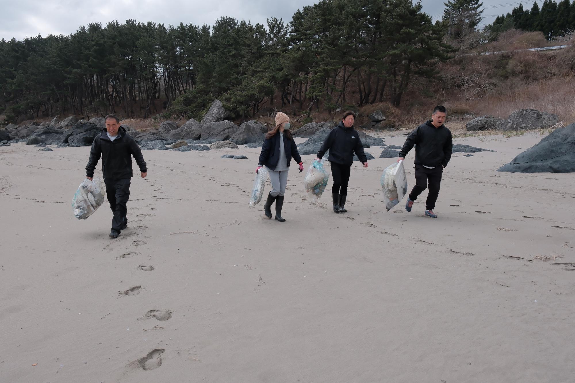砂浜を集めてごみを手に歩く4人の写真