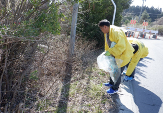 道路脇の草地のごみを拾っている黄色いジャンパーの男性の写真