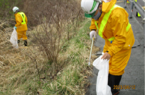 黄色の作業福にヘルメットを着けて道路脇のごみを拾っている二人の写真