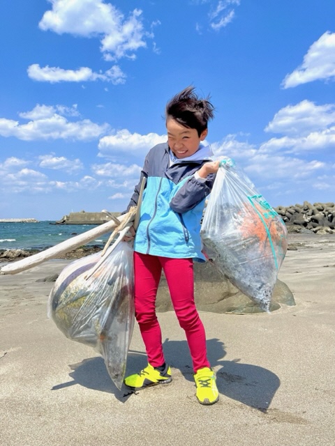青空の砂浜で、重そうなボランティア用ごみ袋を両手に一つづつ持っている児童の写真