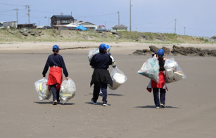 集めたごみ袋を手に、大久喜海岸の砂浜を歩いている3人の生徒の写真