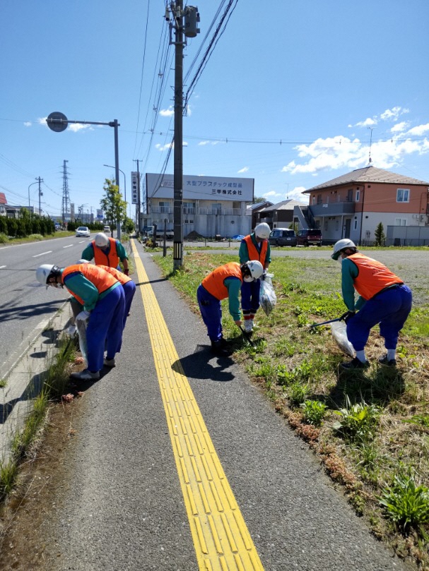 青空の元、同じ作業服にオレンジ色のビブスを身に着け歩道のごみを拾っている5名の写真