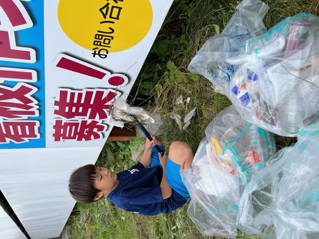 看板の下。ポイ捨てされたごみの入ったボランティア用ごみ袋とトングでごみをつかんでいる男児の写真。